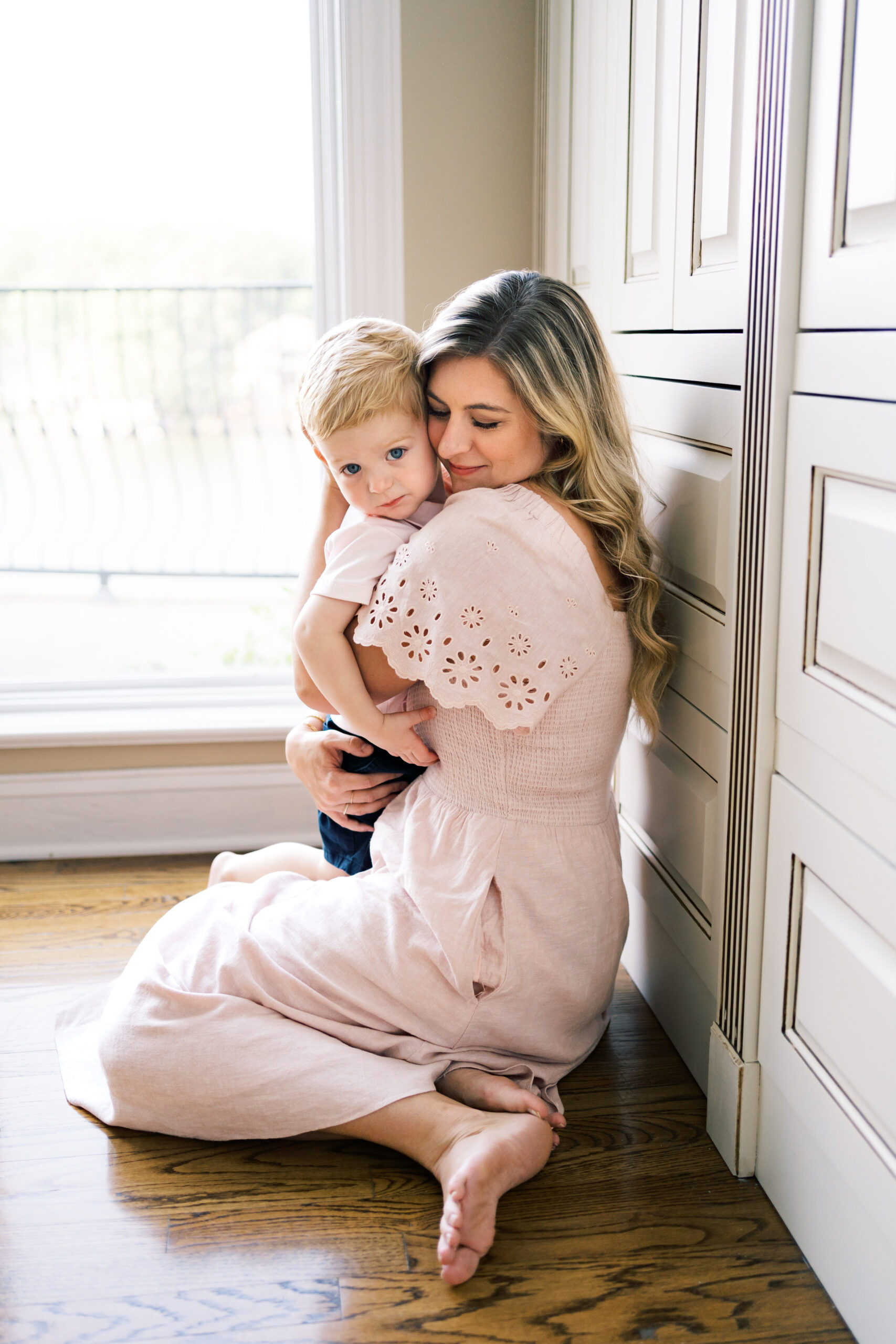 A young boy is hugged by mom while sitting on the floor in a pink dress