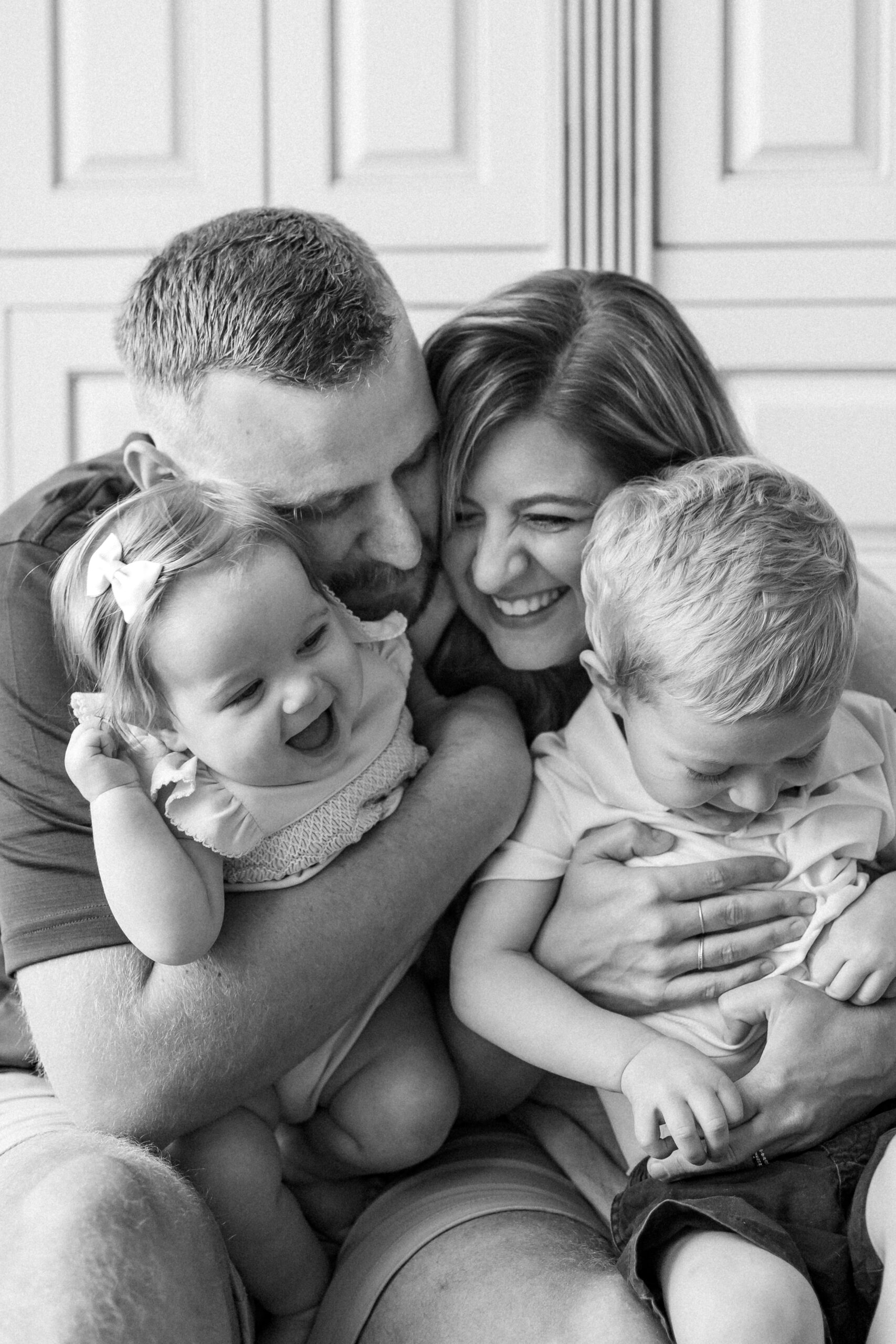 A happy mom and dad hug and tickle their toddler son and daughter in their laps while sitting on a couch