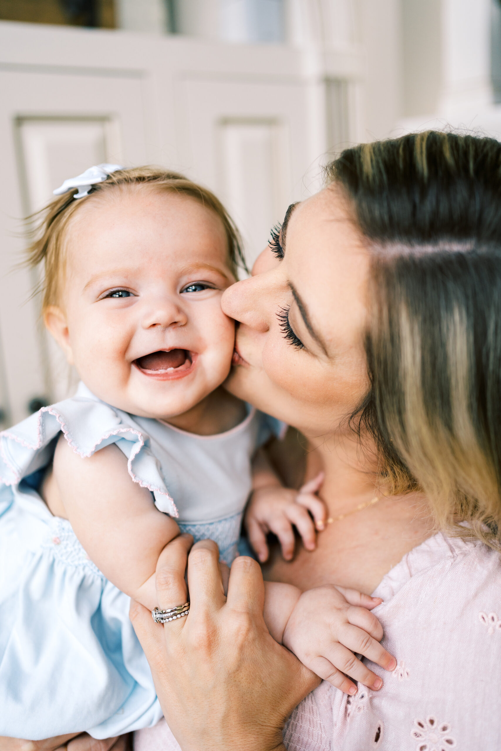 A giggling infant girl in a blue dress laughs while being kissed on the cheek by mom after visiting The Lili Pad