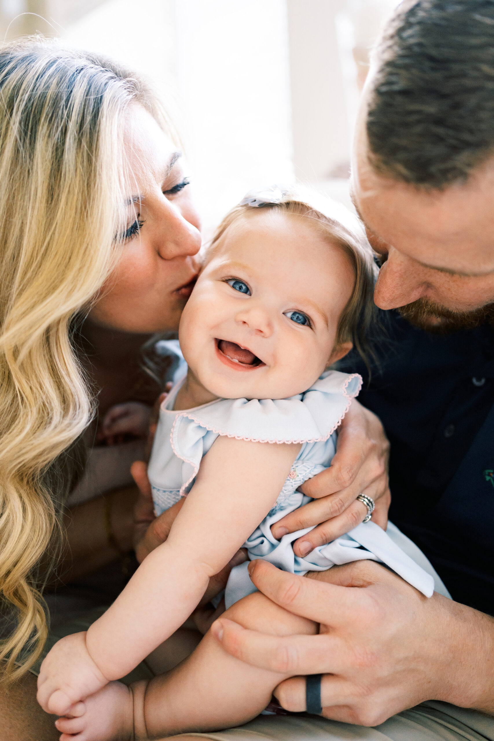 An infant girl sits on a bed and is hugged and kissed by mom and dad after visiting The Lili Pad